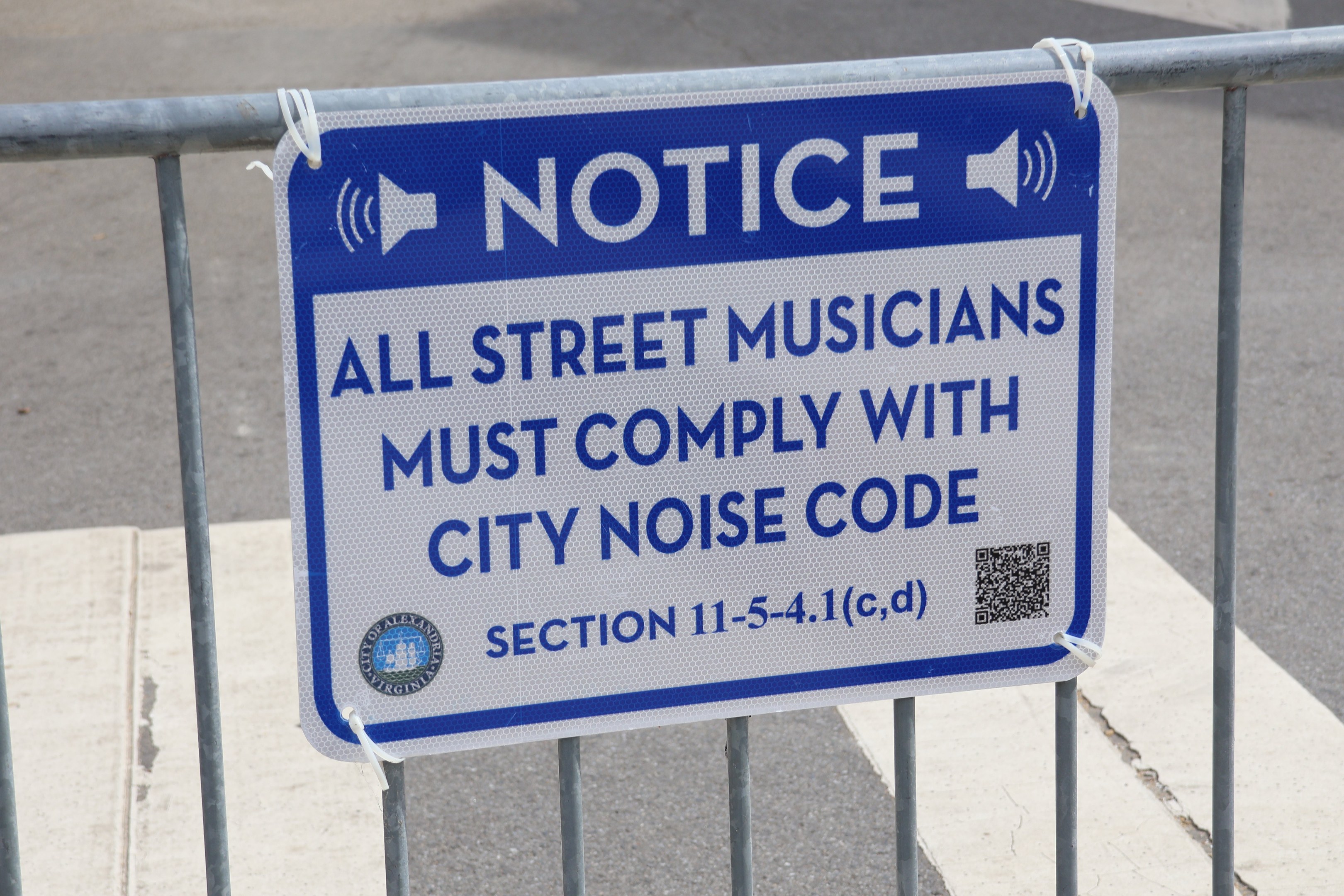 Anschlagtafel an einem Zaun mit einer Tafel, auf der steht "Anschlag: Alle Straßenmusiker müssen die Stadtlärmvorschriften einhalten" und eine Straße im Hintergrund.