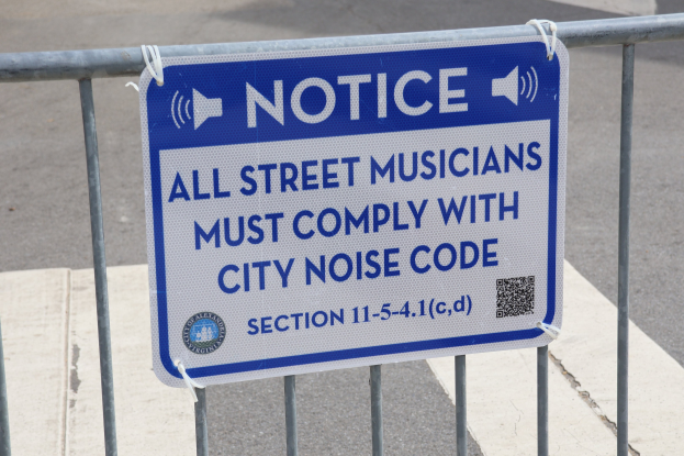 Anschlagtafel an einem Zaun mit einer Tafel, auf der steht "Anschlag: Alle Straßenmusiker müssen die Stadtlärmvorschriften einhalten" und eine Straße im Hintergrund.