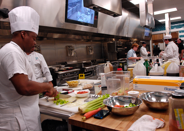 Eine Gruppe von Köchen in einer Küche beim Zubereiten von Speisen auf einem Schneidbrett, mit einem Koch im Vordergrund in einer weißen Schürze und Mütze, der ein Messer hält und von verschiedenen Utensilien, Schüsseln, Gemüse, einem Handy, Flaschen und anderen Gegenständen umgeben ist, mit einem Fernseher und Deckenlampen im Hintergrund.