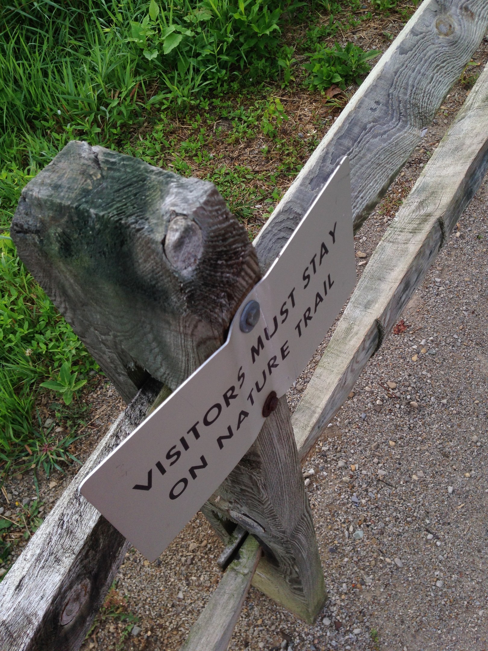 Ein hölzerner Zaun mit einer Tafel, auf der steht "Besucher bleiben auf dem Naturweg", umgeben von grünem Gras und kleinen Steinen.