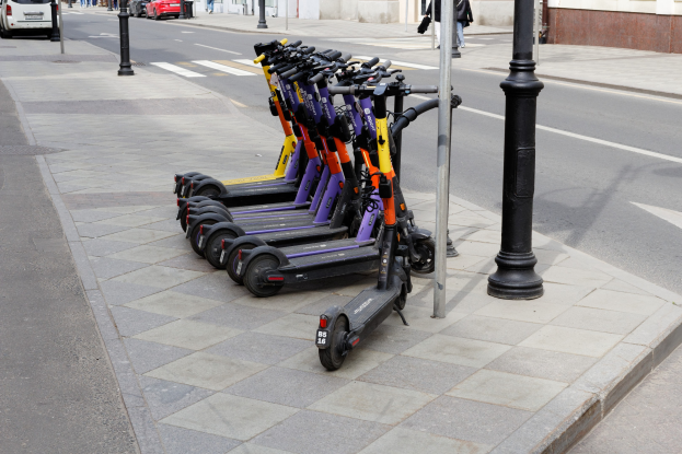 Eine Reihe von Elektro-Scootern, die an einer Straße mit fahrenden Fahrzeugen, Passanten auf dem Gehweg und Gebäuden im Hintergrund geparkt sind.