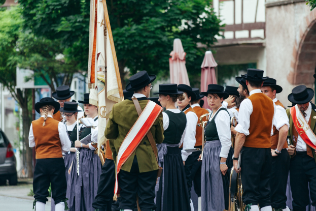 Eine Gruppe von Menschen in traditioneller bayrischer Tracht marschiert auf einer Straße, einige spielen Instrumente und tragen Fahnen, mit Bäumen, Gebäuden und einem Auto im Hintergrund.