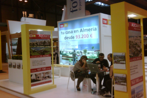 Eine Gruppe von Menschen sitzt um einen Tisch herum und schaut auf einen Laptop, mit verschiedenen Werbeplakaten daneben.