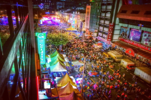 Eine lebhafte Karnevalsstimmung mit einer großen Menschenmenge auf der Straße, umgeben von Zelten, Geschäften, Gebäuden und Bannern, mit zahlreichen Lichtern im Hintergrund.