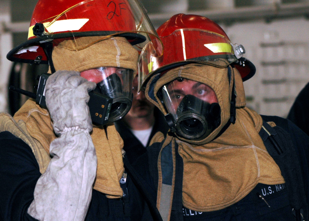 Zwei Feuerwehrleute in Gasmasken und Helmen, einer hält ein Funkgerät in der Hand, mit sichtbaren Rohren an der Wand im Hintergrund.