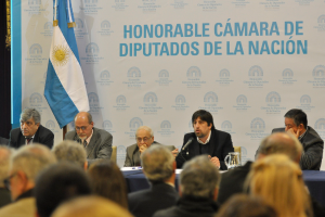 Eine Gruppe von Menschen, die in Reihen von Stühlen sitzen, mit einem zentralen Redner, der ein Mikrofon hält, an einem Tisch mit einem Krug, Papieren und einem Stift, vor einem weißen und blauen Hintergrund mit "ehrenwerte kamara de" darauf geschrieben.