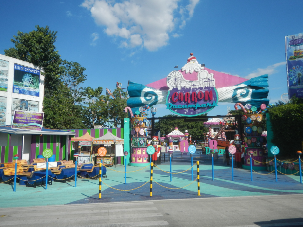 Vergnügungsfahrt am Eingang eines Parks, mit einer Straße, einem Zaun, Buden, Bannern, Bäumen und einem bewölkten Himmel im Hintergrund.