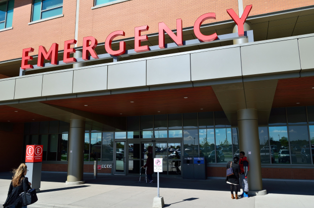 Gruppe von Menschen vor einem Notaufnahmegebäude mit Glaswänden, Säulen und Schildern.