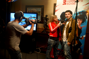 Ein Mann in einem Headset hält eine Videokamera und filmt ein Interview zwischen einem Mann und einer Frau, wobei im Hintergrund ein Fernseher und ein Foto Rahmen zu sehen sind.