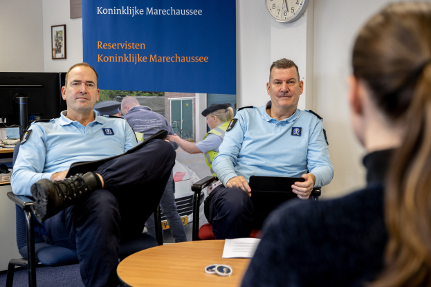 Zwei Polizeibeamte sitzen an einem Tisch mit einer Frau, Papieren und Gegenständen auf dem Tisch, einer Wandtafel und einer Uhr im Hintergrund, einem zweiten Tisch mit einem Monitor, Tastatur, Maus und Gegenständen links daneben und Fotorahmen an der Wand.