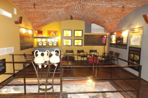 Ein Museumsraum voller Tische, Stühle, einem Geländer, Vasen, Texttafeln, Wandrahmen, Lampen und diversen anderen Ausstellungsgegenständen.