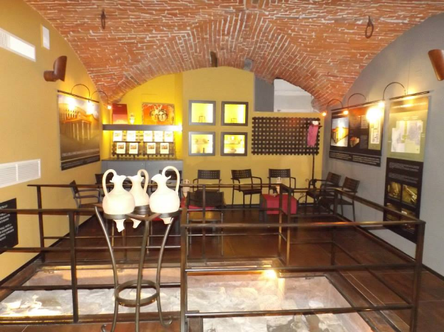 Ein Museumsraum voller Tische, Stühle, einem Geländer, Vasen, Texttafeln, Wandrahmen, Lampen und diversen anderen Ausstellungsgegenständen.