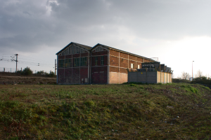 Eine Szene mit Hütten, Transformatoren, Strommasten mit Kabeln, Zäunen, einer Straßenlaterne an einem Laternenmast, Bäumen und einem bewölkten Himmel.
