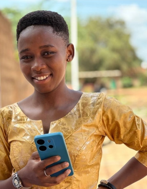 Ein junges Mädchen in einem gelben Kleid hält ein blaues Handy und lächelt vor einem Hintergrund aus Wand, Pfahl, Bäumen und Himmel.