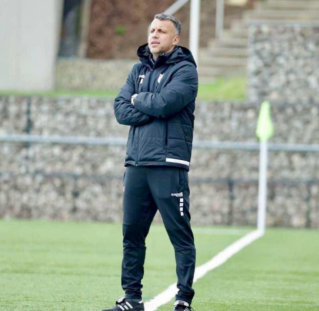 Ein Fussballtrainer, der mit überschlagenen Armen auf einem Feld steht und eine schwarze Jacke und Hose trägt, mit einem Zaun, Treppe und Wand im Hintergrund.