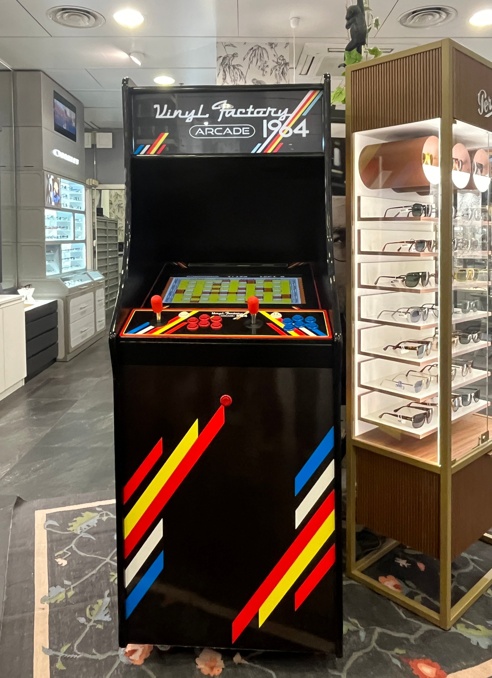 Ein Geschäft mit einem alten Arcade-Automaten in der Mitte, einem Glasvitrine auf der rechten Seite und einer retro-stylischen Beleuchtung mit Schränken, Fernseher und anderen Gegenständen im Hintergrund.