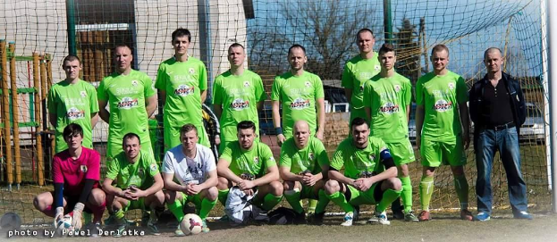Eine Gruppe von Männer in grünen T-Shirts steht auf einem Fussballfeld, einige hocken, mit einem Ball vor ihnen in der Nähe eines Torpfostens, Bäume und Gebäude im Hintergrund.