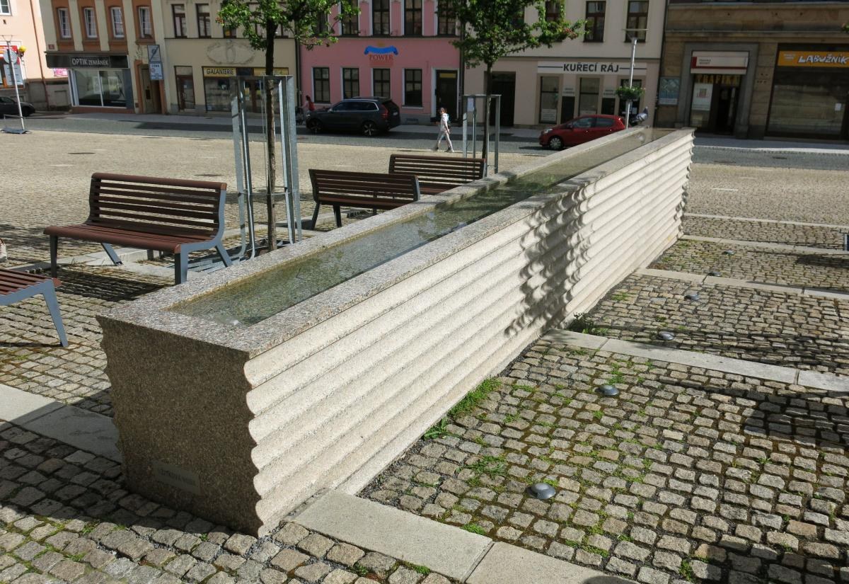 Stadtplatz mit einem zentralen Brunnen, umgeben von Bänken, Bäumen und Pfählen, mit Fahrzeugen auf der Straße und Menschen im Hintergrund bei Gebäuden mit Fenstern und Namensschildern.