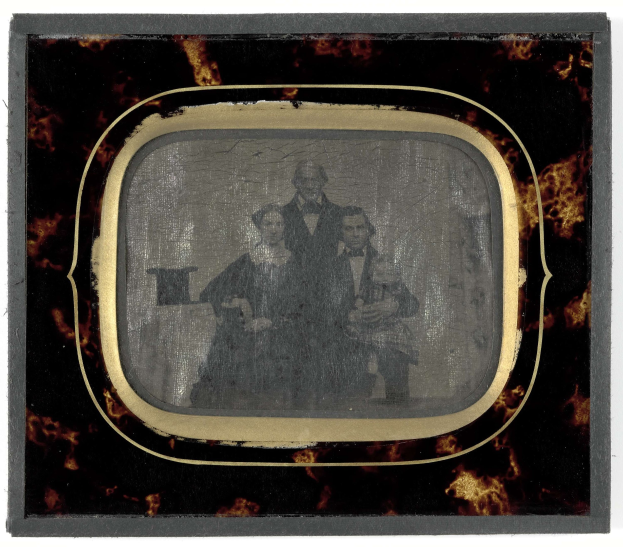 Schwarze-Weiß-Foto eines Mannes in Anzug und Krawatte und einer Frau in unbekannter Kleidung, die vor einem alten Fernseher stehen, beide mit ernsten Gesichtern.