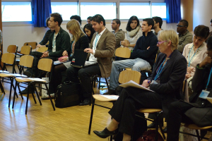 Mehrere Personen auf Stühlen sitzend, mit ID-Karten, wobei ein Mann einen Laptop und eine Frau einen Stift hält, umgeben von leeren Stühlen, Fenstern und Vorhängen auf einem Boden mit einer Tasche in der Nähe.