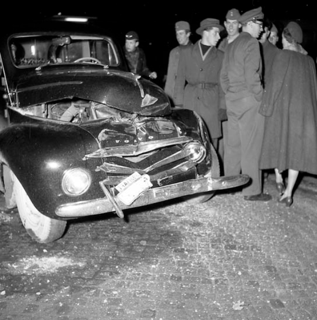 Ein Schwarz-Weiß-Bild eines beschädigten Autos am Straßenrand, umgeben von einer Gruppe besorgter Menschen.