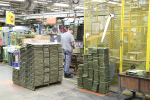 Ein Mann in einer Mütze und T-Shirt steht in einer Fabrik, arbeitet in der Nähe von Maschinen auf dem Boden und an der Decke, mit Lüftern und Beleuchtung darüber.