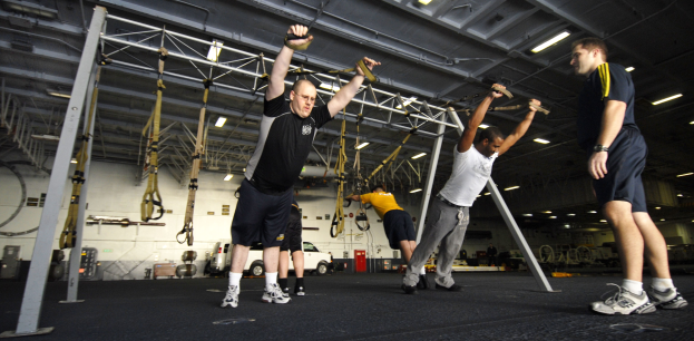 Mannergruppe beim Pull-up an Metallstangen in einem Fitnessstudio mit Deckenbeleuchtung und diversen Gegenständen im Hintergrund, mittig ein Mann in Schuhen.