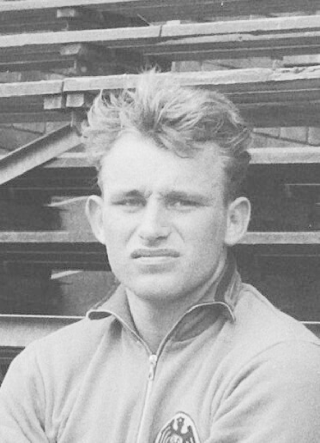 Schwarzes und weißes Foto eines Mannes in einer Jacke, der vor gestapelten Holzplanken steht, der angeblich ein legendärer deutscher Fußballspieler ist.