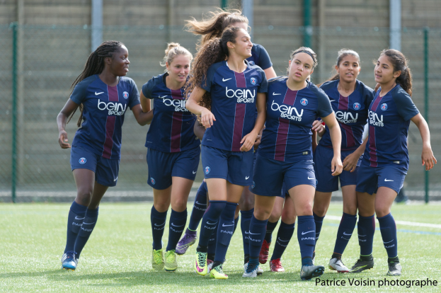 Eine Gruppe junger Frauen, die auf einem Rasenfeld Fußball spielen, mit Maschendraht und einer Wand im Hintergrund, beschriftet mit "Paris Saint-Germain Frauenfußball" in der rechten unteren Ecke.