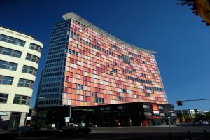 Hohe Gebäude mit parkenden Autos in einer Parkplatzfläche und ein Verkehrsampelmast auf der rechten unteren Seite.