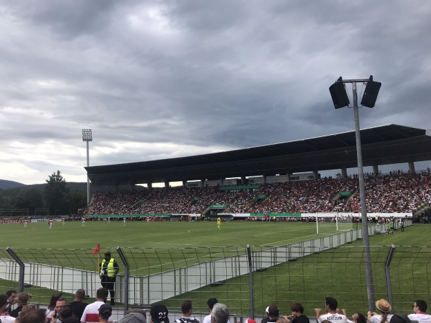 Große Zuschauermenge bei einem Fußballspiel in einem eingezäunten Stadion mit beleuchtetem Feld, Bäumen, Hügeln und bewölktem Himmel im Hintergrund.