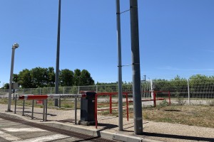 Rotes und weißes Schild an einem Bahnhof umgeben von einem Metallzaun, Pfählen, Bäumen, Pflanzen, Gras, einem Straßenpfahl, Drähten und einem bewölkten Himmel, mit einer Absperrung im Vordergrund.