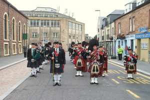 Eine Gruppe von Menschen, einige stehen auf der Straße und andere spielen Musikinstrumente, mit Gebäuden, Plakaten, Polen, Bäumen und Reifen im Hintergrund.