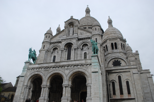Ein Denkmal steht in der Mitte des Bildes, mit Häusern und einem Baum in der unteren linken Ecke.