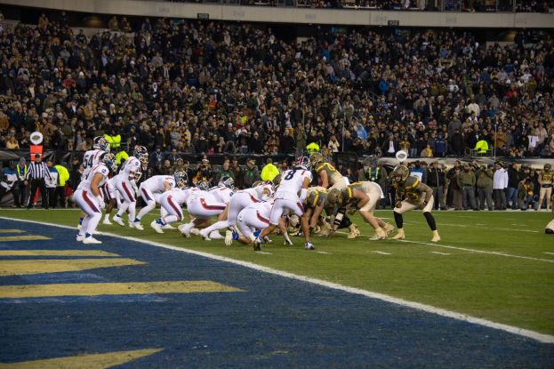 Ein Fußballspiel im Gange mit Spielern in Sportbekleidung und Helmen auf dem Boden, von einem Stadionplatz aus betrachtet mit Zuschauern, Geländern und Werbetafeln im Hintergrund.
