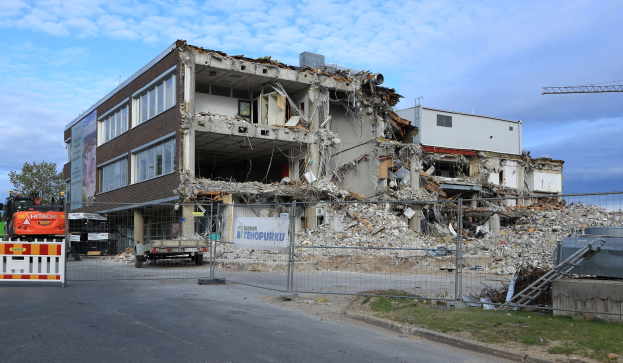 Großes Gebäude in der Mitte einer Straße abgerissen, umgeben von einem Metallzaun, Fahrzeugen, Gras, einem Schild, einer Fahne, Pfählen, einem Kran, Bäumen und einem bewölkten Himmel.