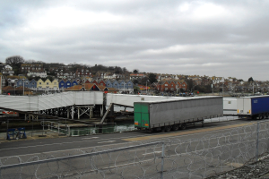 Ein Sattelschlepper fährt auf einer Straße neben einem Stacheldrahtzaun, mit einer Brücke, Gebäuden, Bäumen und einem bewölkten Himmel im Hintergrund.