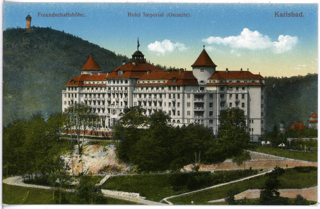Eine alte Postkarte des Hotel Imperial in Karlsbad, Deutschland, zeigt ein großes Gebäude mit Fenstern, umgeben von Bäumen, Pflanzen, Gras, einem Pfad, einer Gruppe von Bäumen, Hügeln und einem bewölkten Himmel, mit Text oben.