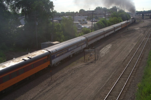 Ein Zug auf Schienen mit B√§umen, Stromm√§sten mit Kabeln, Autos und Geb√§uden links, und eine Br√ľcke vorne, die Rauch ausst√∂√üt.