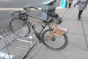 Ein grünes und braunes Fahrrad steht auf der Straße neben einem Müllcontainer, mit einigen Gegenständen daran befestigt, während Fahrzeuge auf der Straße fahren und Fußgänger im Hintergrund zu sehen sind.