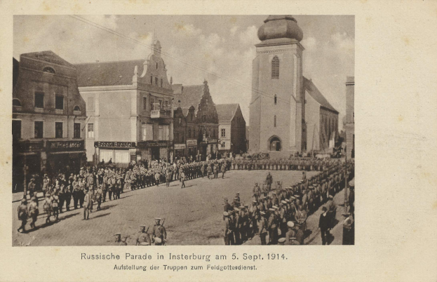 Ein Schwarz-Weiß-Foto eines Umzugs in Insterburg am 5. September 1914 mit vielen Menschen, Gebäuden im Hintergrund und Text unten auf dem Bild.
