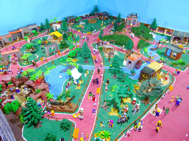 Ein Miniaturmodell eines Zoos mit Besuchern, umgeben von Bäumen, Häusern, Pflanzen, Gras, einem Zaun und einer Wand mit Text unten.