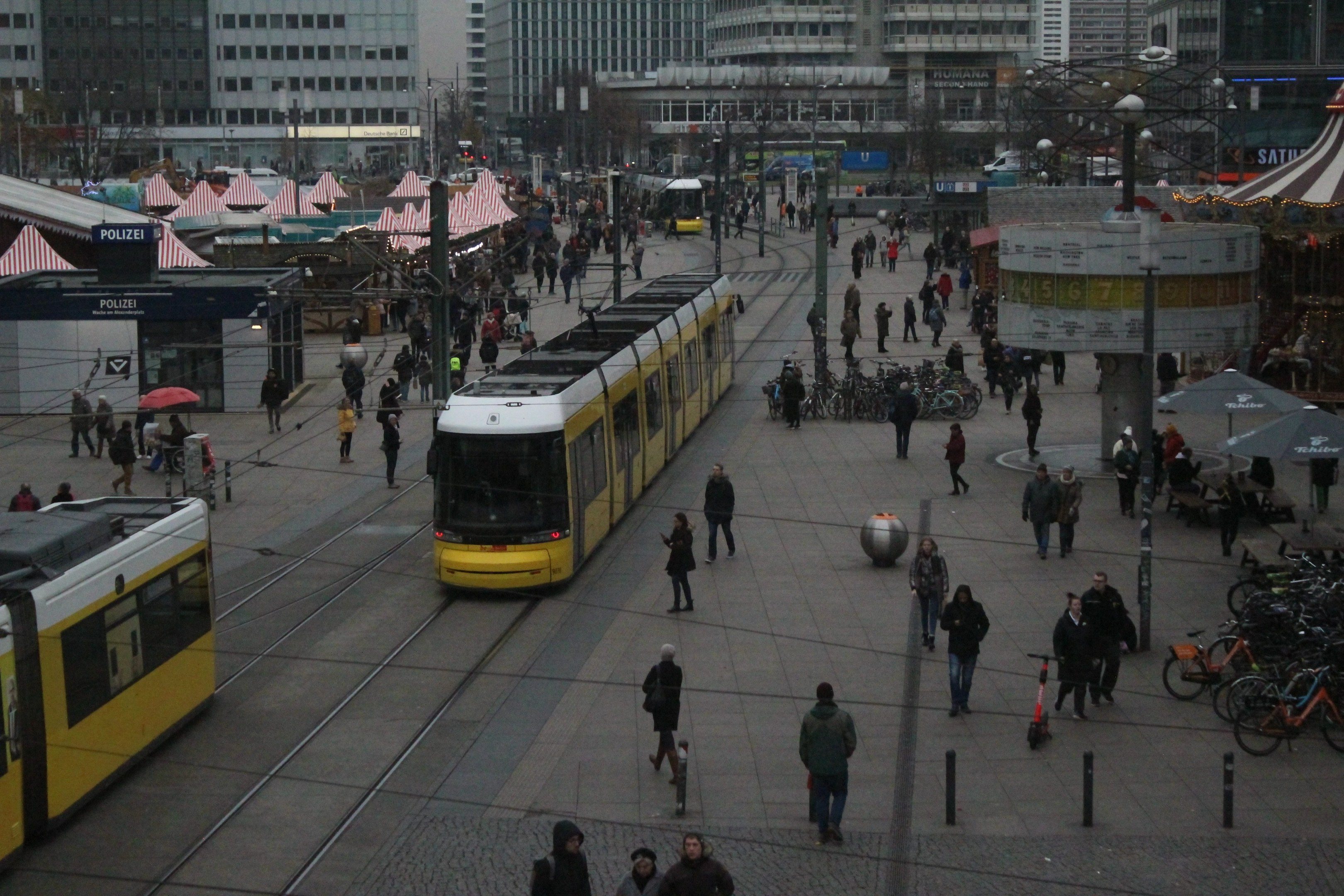 Eine belebte Stadtstraße mit Fußgängern, Radfahrern, Regenschirmen, Straßenlaternen, Zelten, Gebäuden, Bäumen und einem klaren blauen Himmel.