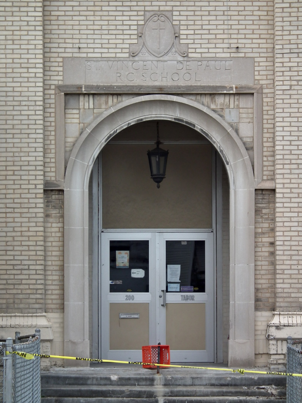 Eingang eines Backsteingebäudes mit gelbem Absperrband, Treppe mit Maschendrahtzaun, eine Tür mit Papieren, eine hängende Lampe und ein Name an der Wand.