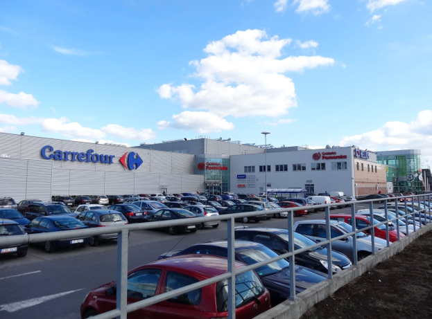 Großes Autohaus mit zahlreichen Fahrzeugen auf dem Parkplatz, umgeben von Gebäuden und Straßeninfrastruktur unter einem bewölkten Himmel.
