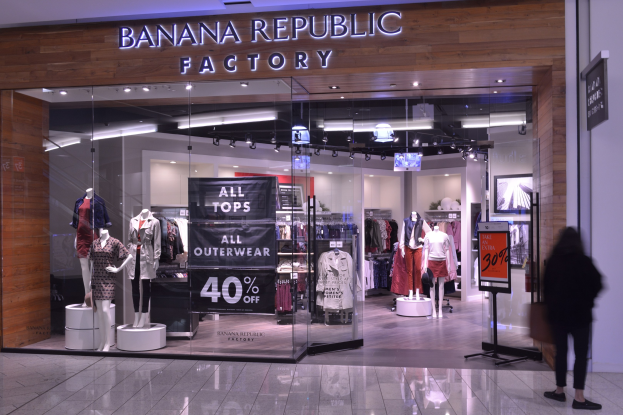 Außenansicht eines Banana Republic Factory Stores in Singapur mit Schaufensterpuppen in der neuesten Mode, hängenden Kleidern, Informationsschildern, Deckenleuchten und einer Person im Inneren.