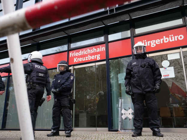 Eine Gruppe von Polizisten in Helmen und Handschuhen steht vor einem Glasgebäude, mit einem Pfahl links und Schildern im Hintergrund, die "Friedrichshain Kreuzberg" und Papiere an den Glaswänden kleben.