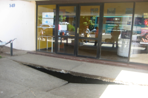Eine gebrochene Oberfläche im Vordergrund mit einer Glastür, Wand und einem Pfahl im Hintergrund, die sich bewegende Fahrzeuge und einige stehende Personen spiegeln.