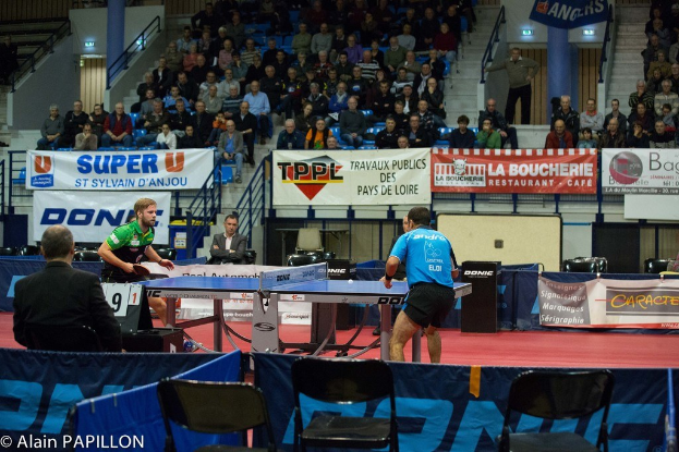 Indoor-Tischtennis-Match mit zwei Spielern und einigen Zuschauern, die auf Stühlen sitzen und Werbeplakate in einer Stadionatmosphäre.