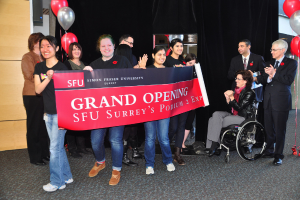 Eine Gruppe von Frauen hält ein Banner hoch, um ein Event zu bewerben, daneben stehen zwei klatschende Männer und eine Frau im Rollstuhl, die begeistert aussieht, vor einem schwarzen Vorhang und Ballons.